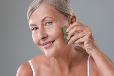 Photo of Beautiful woman doing facial massage with gua sha tool on grey background, closeup