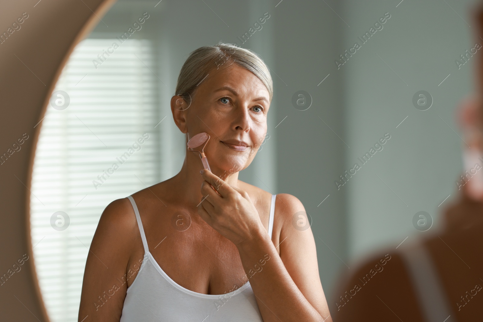 Photo of Beautiful woman doing facial massage with roller near mirror at home