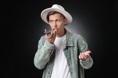 Young man recording voice message via smartphone on dark background