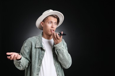 Photo of Young man recording voice message via smartphone on dark background, space for text