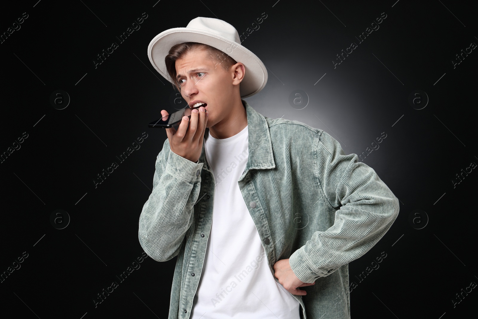 Photo of Young man recording voice message via smartphone on dark background