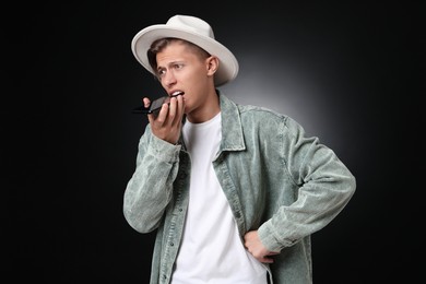 Young man recording voice message via smartphone on dark background