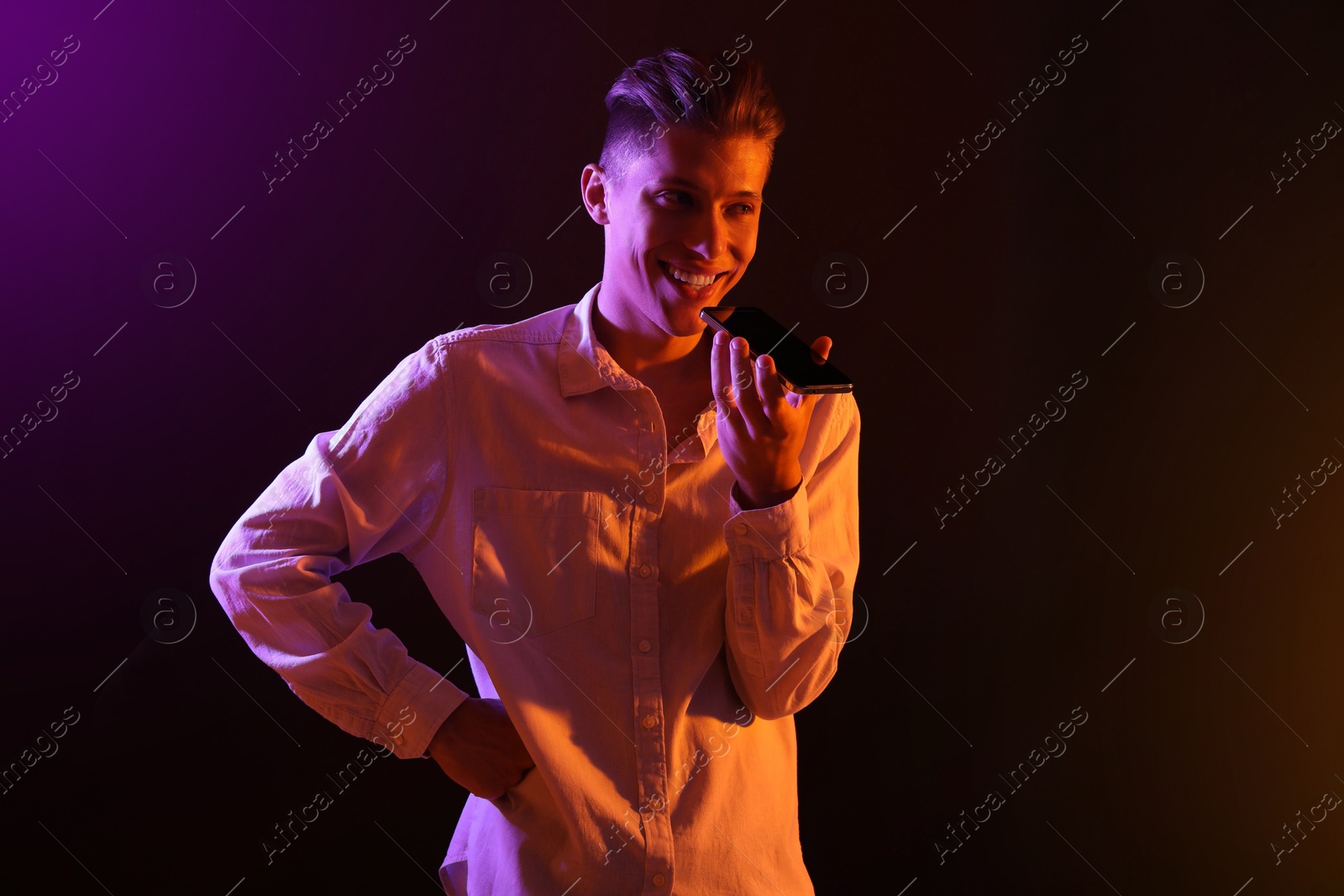 Photo of Young man recording voice message via smartphone on dark background with neon lights