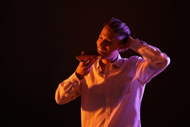 Young man recording voice message via smartphone on dark background with neon lights, space for text