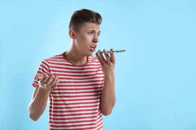 Young man recording voice message via smartphone on light blue background, space for text