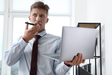 Photo of Young man with laptop recording voice message via smartphone in office