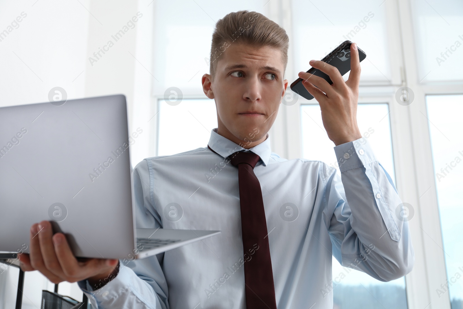 Photo of Young man with laptop listening to voice message via smartphone in office