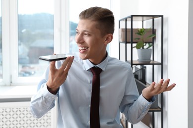 Photo of Young man recording voice message via smartphone in office