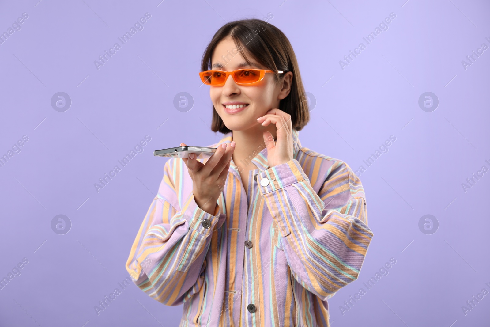 Photo of Beautiful woman in sunglasses recording voice message via smartphone on violet background