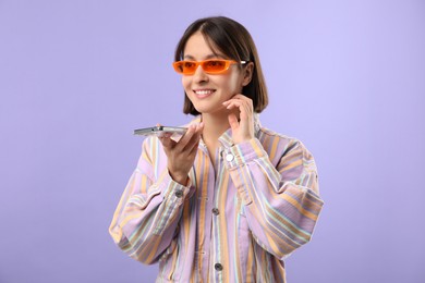 Beautiful woman in sunglasses recording voice message via smartphone on violet background