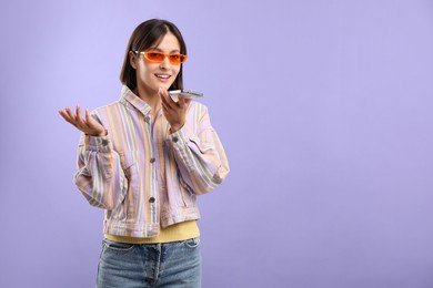 Beautiful woman in sunglasses recording voice message via smartphone on violet background, space for text