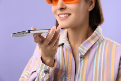 Photo of Woman in sunglasses recording voice message via smartphone on violet background, closeup