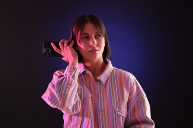 Beautiful woman listening to voice message via smartphone on dark background with neon lights