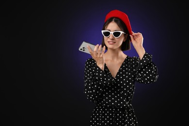 Stylish woman recording voice message via smartphone on dark background, space for text