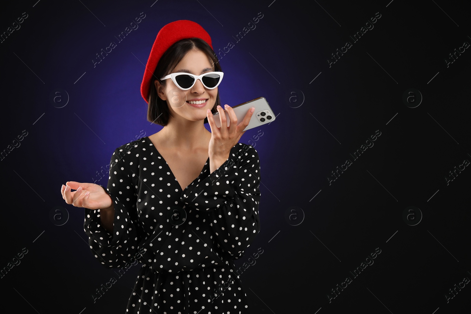 Photo of Stylish woman recording voice message via smartphone on dark background, space for text