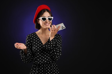 Stylish woman recording voice message via smartphone on dark background, space for text