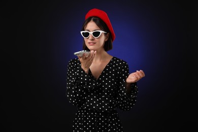 Stylish woman recording voice message via smartphone on dark background