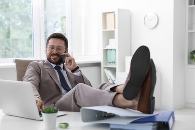 Photo of Smiling businessman talking by smartphone and holding legs on table in office