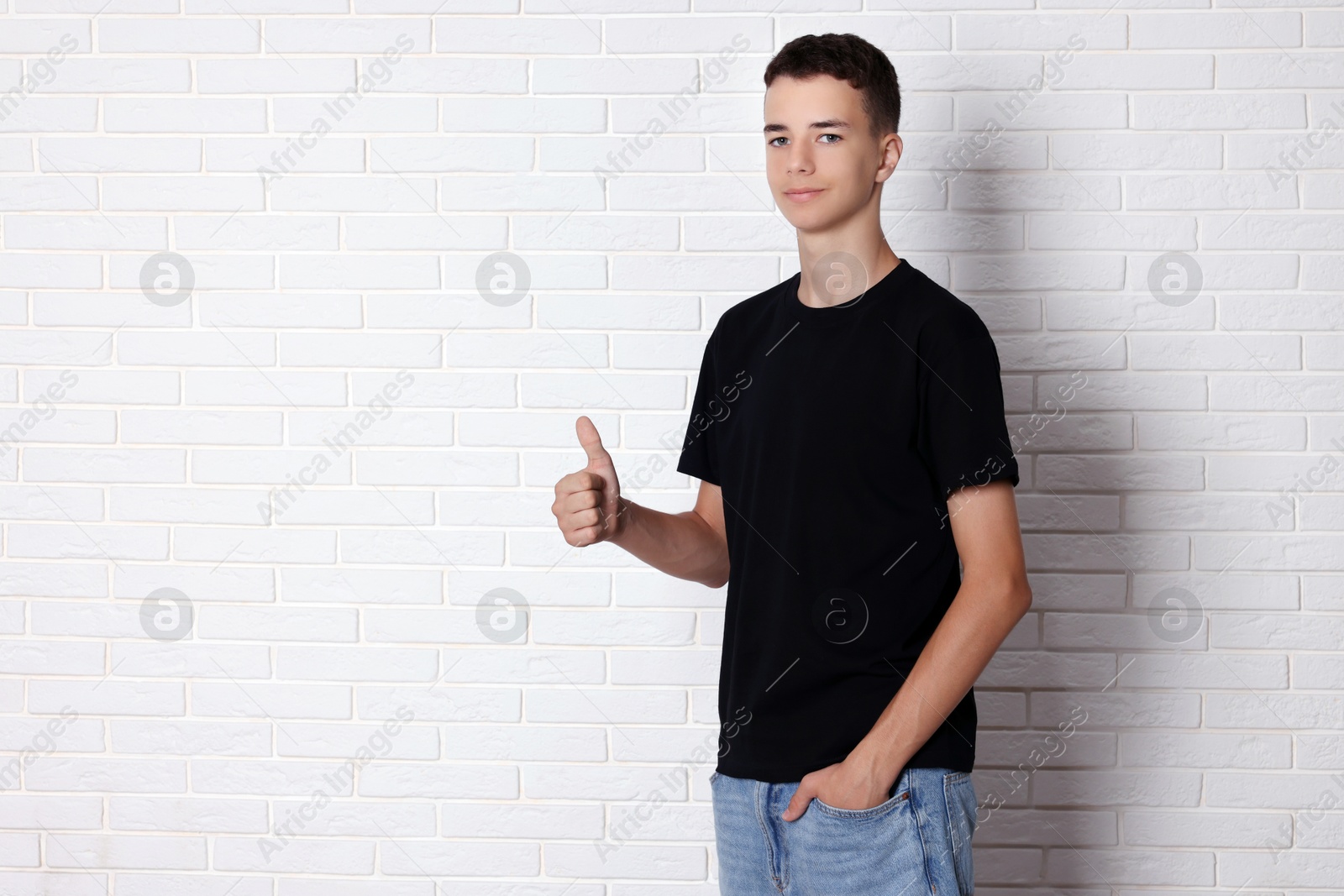 Photo of Teenage boy wearing black t-shirt and showing thumbs up near white brick wall, space for text