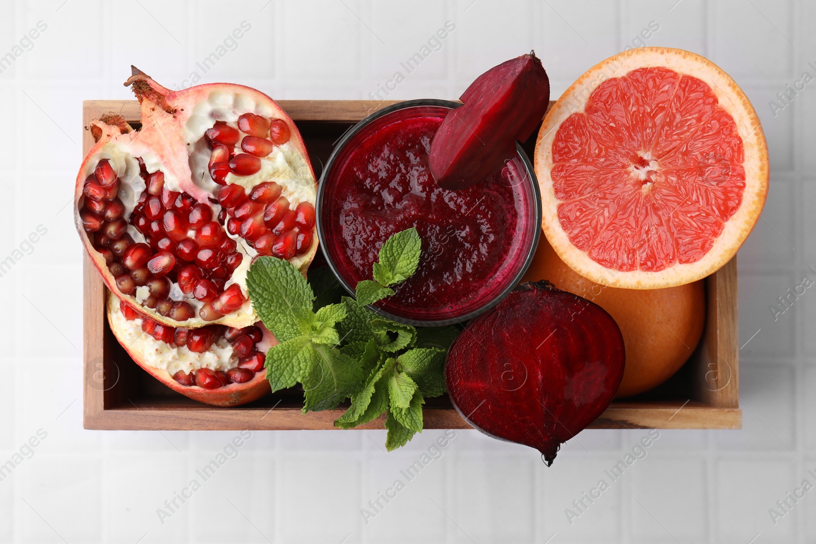 Photo of Glass of tasty beetroot smoothie, fresh vegetables, fruits and mint in wooden box on white tiled table, top view. Vegan drink