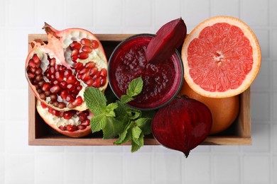 Photo of Glass of tasty beetroot smoothie, fresh vegetables, fruits and mint in wooden box on white tiled table, top view. Vegan drink