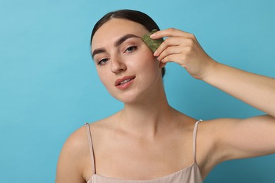 Photo of Beautiful young woman doing facial massage with gua sha tool on light blue background