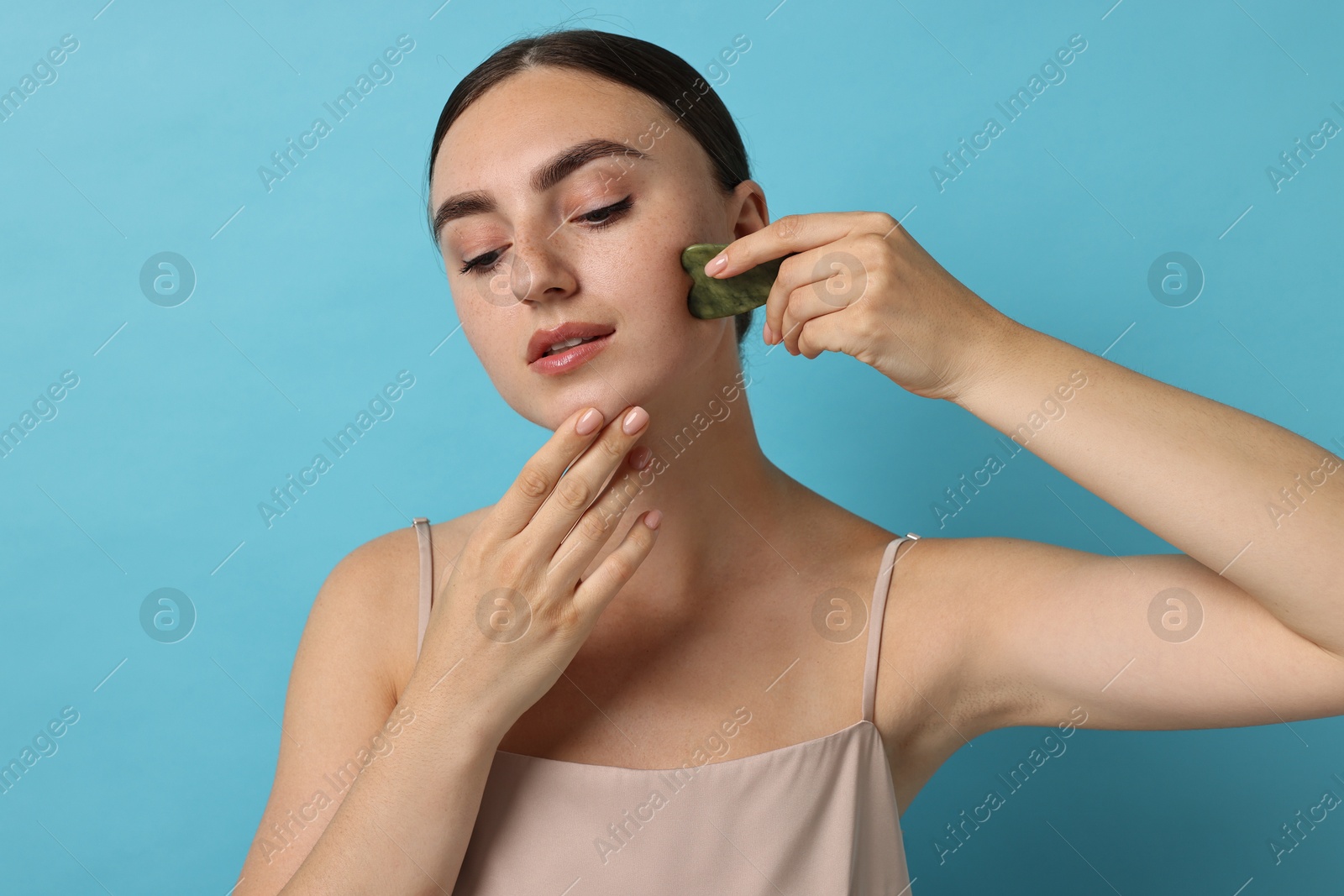 Photo of Beautiful young woman doing facial massage with gua sha tool on light blue background