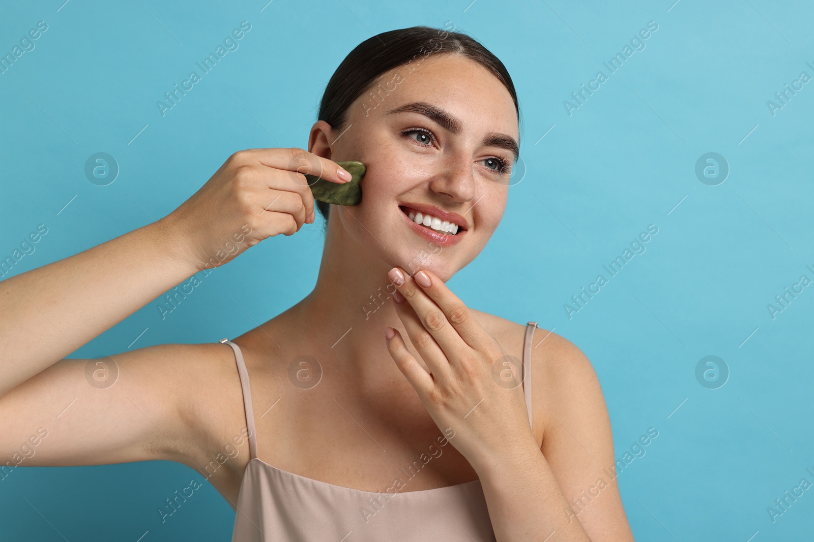 Photo of Beautiful young woman doing facial massage with gua sha tool on light blue background