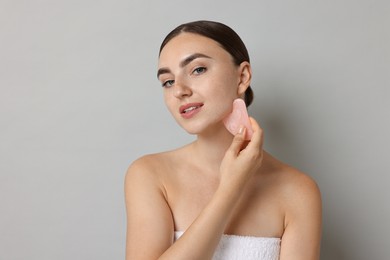 Photo of Beautiful young woman doing facial massage with gua sha tool on grey background