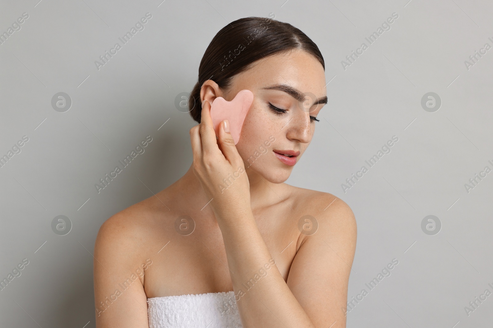 Photo of Beautiful young woman doing facial massage with gua sha tool on grey background