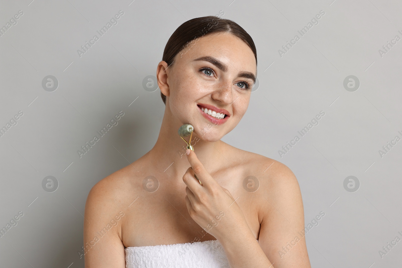 Photo of Beautiful young woman doing facial massage with roller on grey background