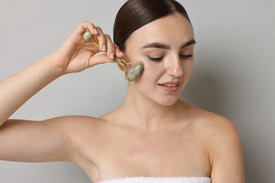 Beautiful young woman doing facial massage with roller on grey background