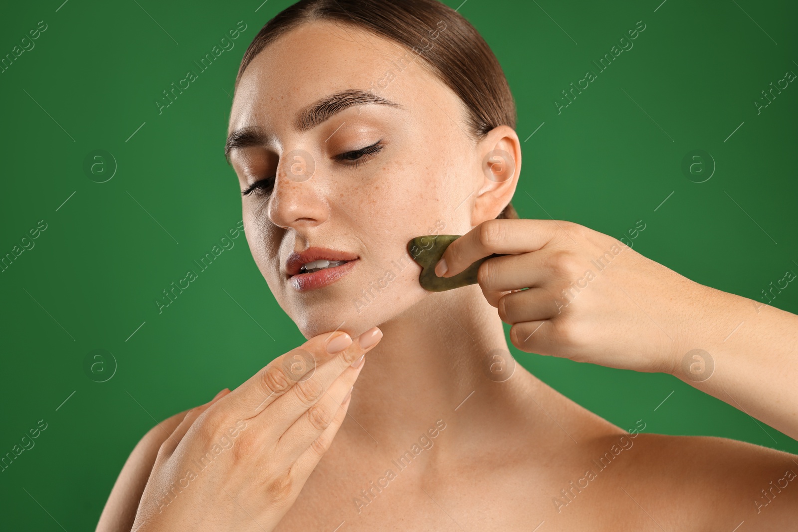 Photo of Beautiful young woman doing facial massage with gua sha tool on green background