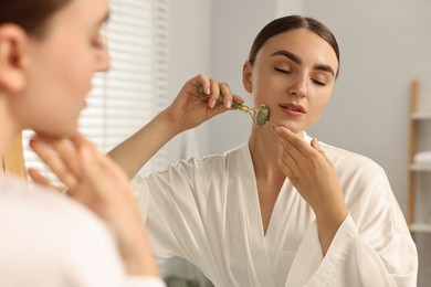 Photo of Beautiful young woman doing facial massage with roller near mirror at home