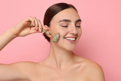 Beautiful young woman doing facial massage with roller on pink background