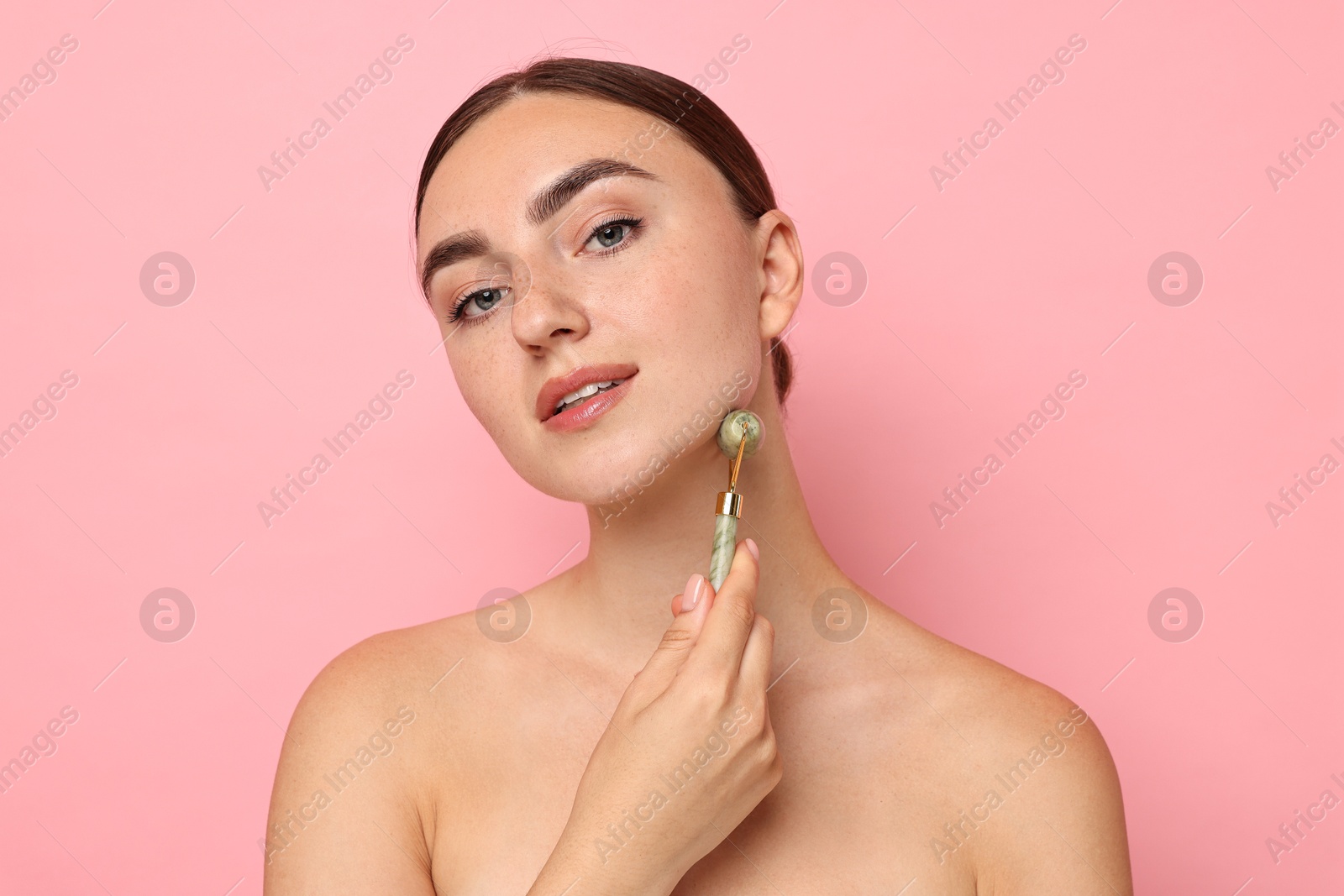 Photo of Beautiful young woman doing facial massage with roller on pink background