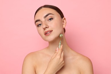 Beautiful young woman doing facial massage with roller on pink background