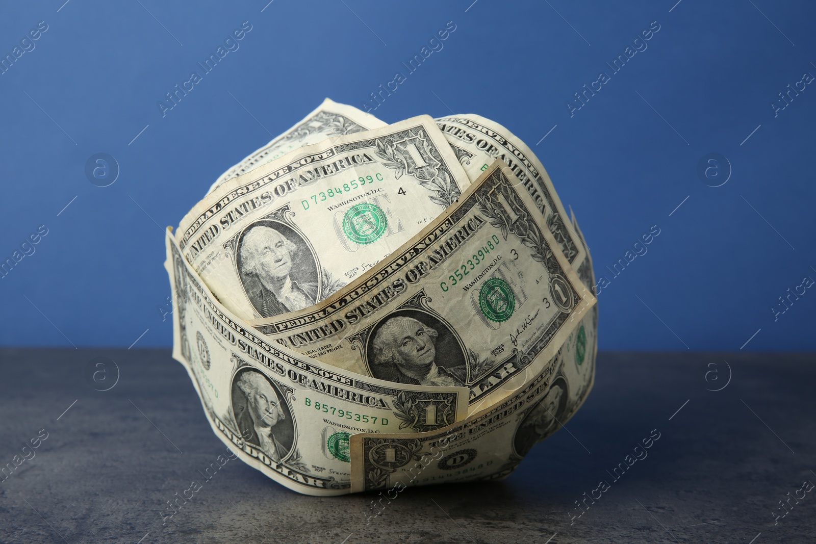 Photo of Ball made of dollar banknotes on grey textured table against blue background, closeup