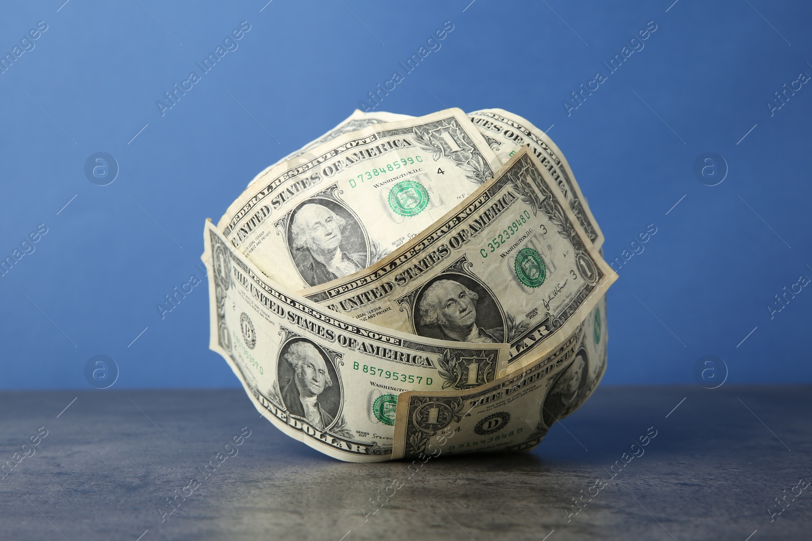 Photo of Ball made of dollar banknotes on grey textured table against blue background, closeup