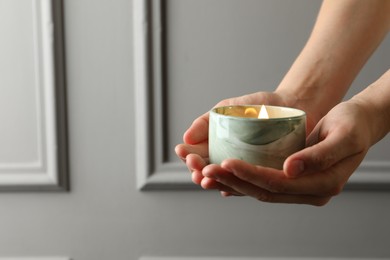 Woman holding burning candle against grey wall, closeup. Space for text