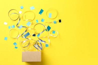 Photo of Box with confetti and party streamers on yellow background, flat lay. Space for text