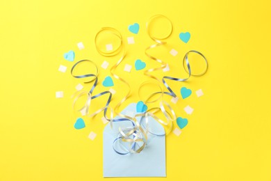 Envelope with confetti and party streamers on yellow background, flat lay