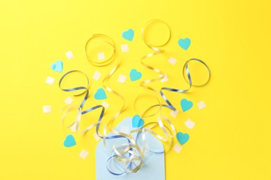 Envelope with confetti and party streamers on yellow background, flat lay