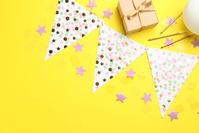 Confetti, garland, candles and gift box on yellow background, flat lay. Space for text