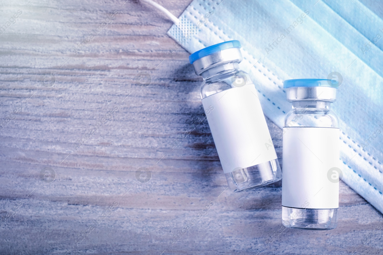 Image of Medication in glass vials with blank labels and medical mask on wooden table, flat lay. Space for text