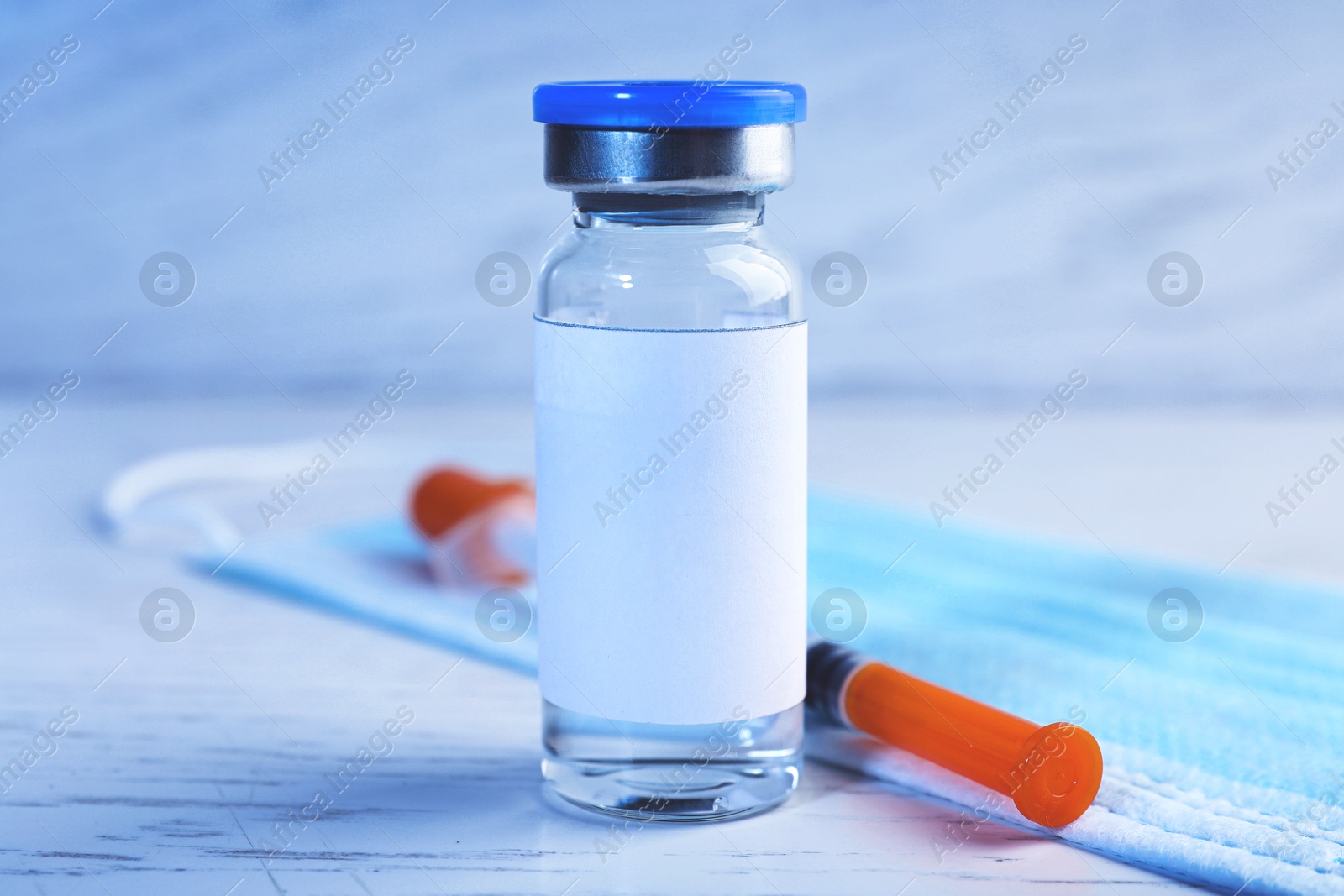 Image of Medication in glass vial with blank label, medical mask and syringe on white wooden table, closeup