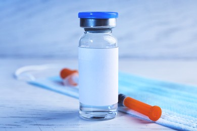 Medication in glass vial with blank label, medical mask and syringe on white wooden table, closeup