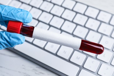 Laboratory worker holding test tube with blood sample and blank label over computer keyboard, closeup. Medical analysis