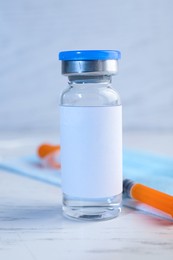 Medication in glass vial with blank label, medical mask and syringe on white wooden table, closeup