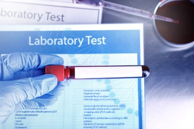 Laboratory worker holding test tube with blood sample and blank label over table, closeup. Medical analysis